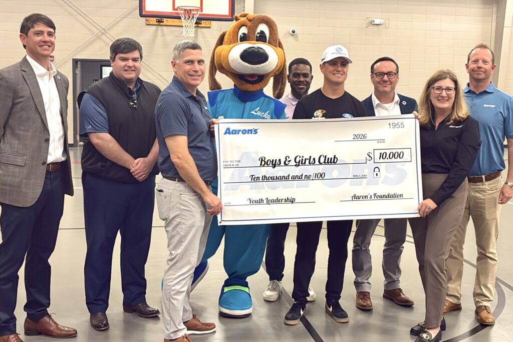 A large group of company and nonprofit leaders – with Aaron’s mascot Lucky Dog at the center – stand together smiling and holding up a giant $10k check from Aaron’s to the organization.