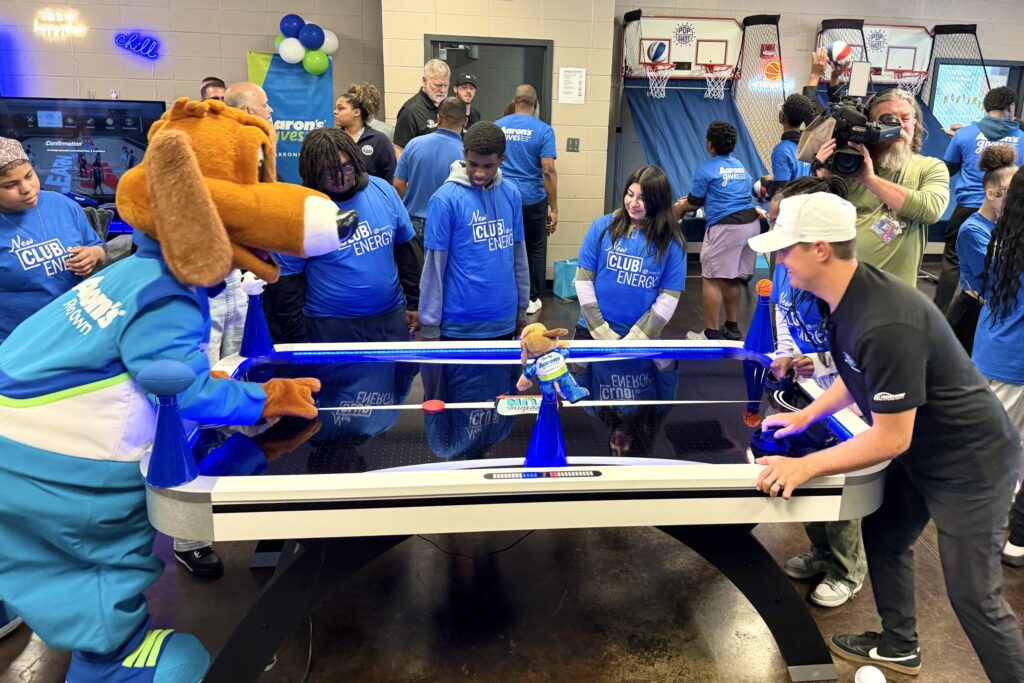The Lucky Dog mascot and Zane Smith play air hockey at opposite ends of the table as Club members and others watch the match.