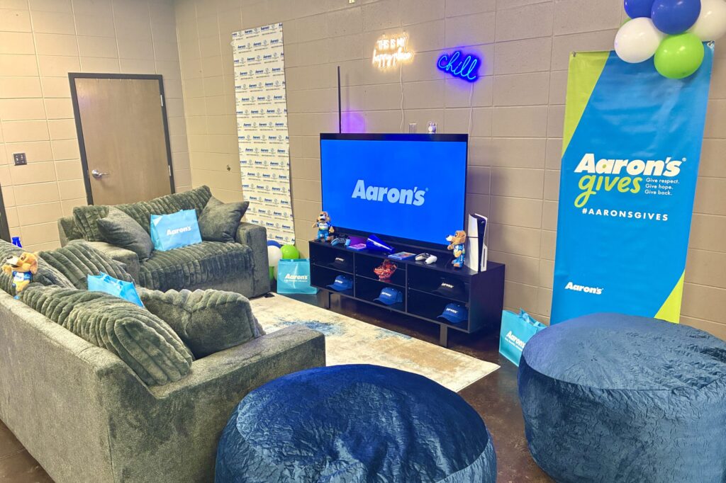 Two big blue sofas and two velvet stools face a big-screen TV with the Aaron’s logo, flanked by two Aaron’s banners.