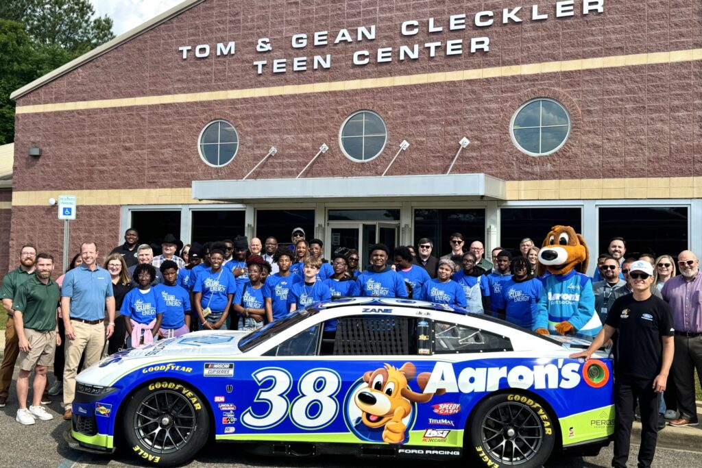 A large group of Aaron’s and Boys & Girls Clubs of Central Alabama representatives, along with company mascot Lucky Dog and teen members of the Club, stand smiling in front of the Tom and Gean Cleckler Teen Center and behind a white racecar. Driver Zane Smith stands alongside the car, also smiling.