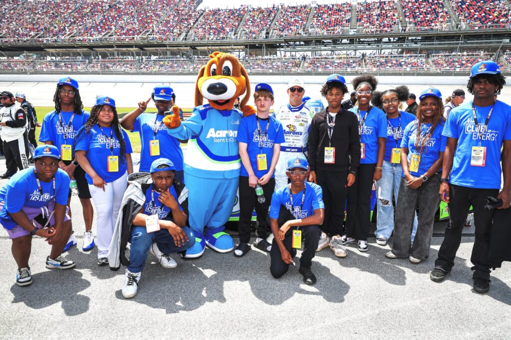 group of teenagers, Aaron’s Lucky Dog mascot, and NASCAR driver Zane Smith stand smiling by the Talladega Superspeedway racetrack with a crowd in the grandstand behind them.