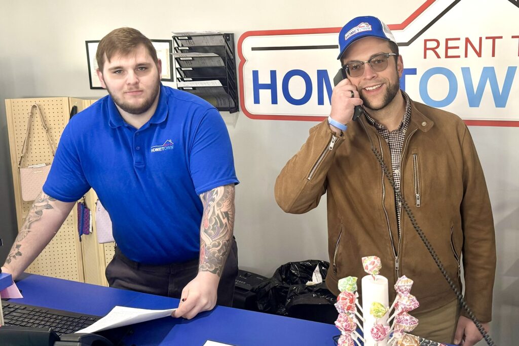 Adam Harvey stands behind the counter at the Hometown RTO store he manages, while J.P. Bryant stands beside him on the phone and smiling.