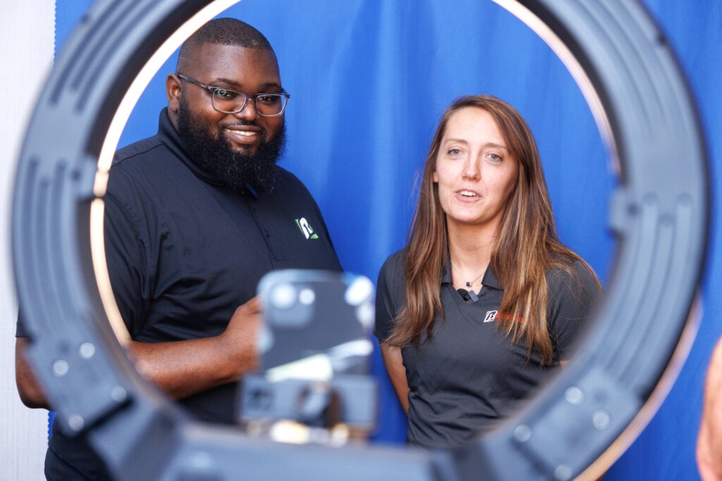 A man and woman wearing Rent One shirts gaze and smile through the encircling frame of a ring light. 
