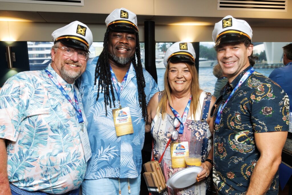 Three men and a woman wearing tropical shirts and boat captain caps smile with blue water behind them. 
 