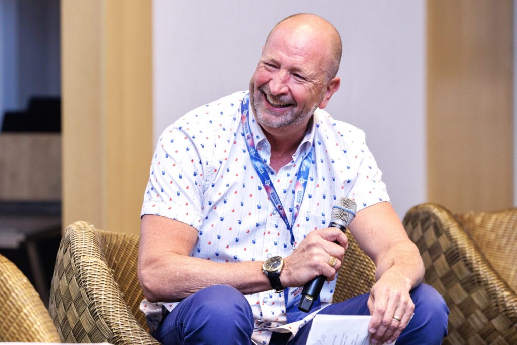 Trent Agin sits in a chair, holding a microphone, and laughing heartily. 
 
 