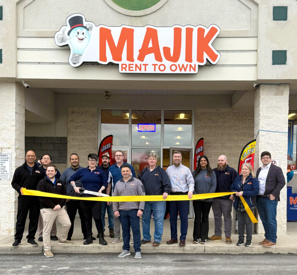 A group of men and women stand outside a Majik Rent-To-Own storefront holding a large yellow ribbon during a grand opening celebration.