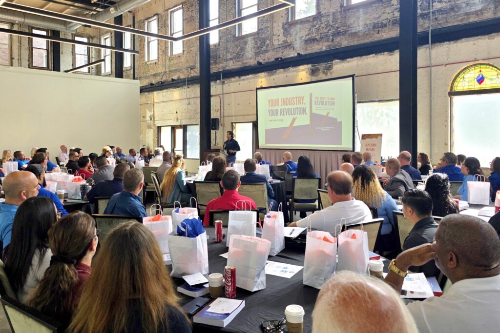Dozens of people seated at tables listen intently to Smitherman as he presents from a stage with a screen behind him reading “Your Industry, Your Revolution.”