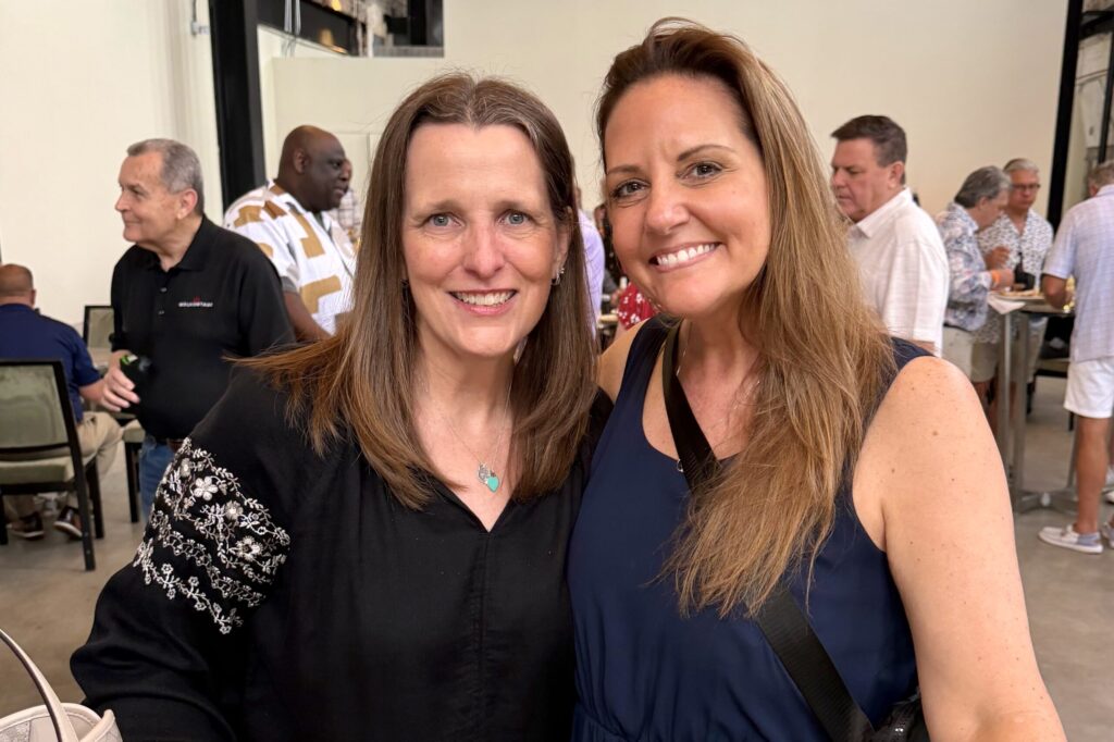 Two women stand together and smile brightly with a crowd behind them.