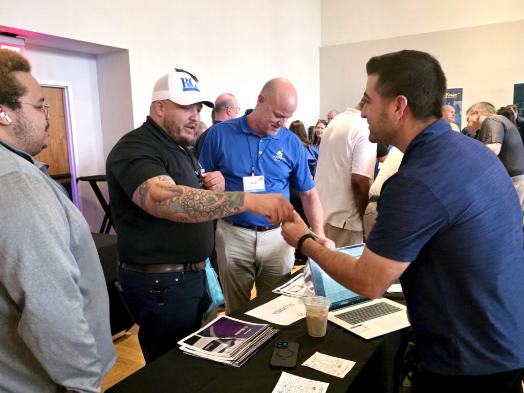 Two men interact over a table holding a laptop and informational paperwork while a crowd of others move about around them.