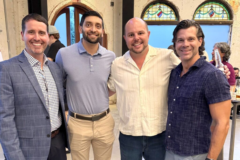 Four men stand side-by-side with arms around each other, smiling.