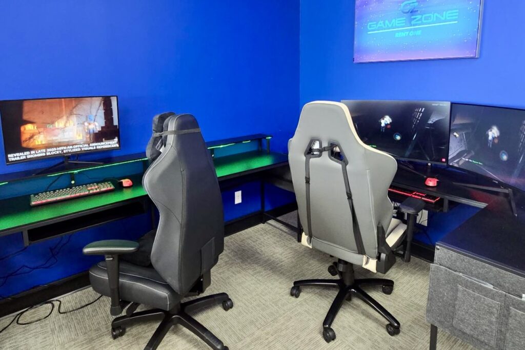 Two gaming stations with dark tables, ergonomic chairs, colorful monitors, and gaming equipment are positioned beneath an LCD screen that reads “Game Zone – Rent One.” 