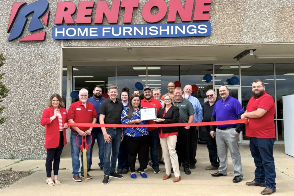 A large group of Rent One leaders and team members stand smiling in front of a Rent One storefront, with two people holding up a long red ribbon and one holding a giant pair of scissors. 