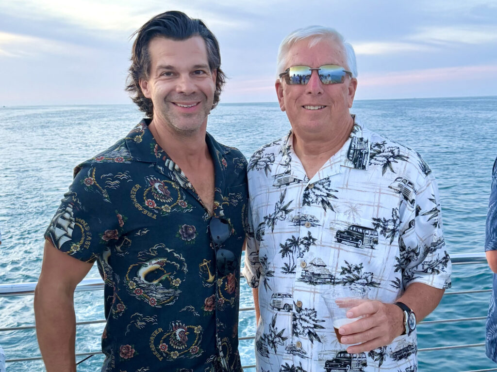 Two men wearing Hawaiian-style shirts smile with the ocean and a dusky sky behind them.