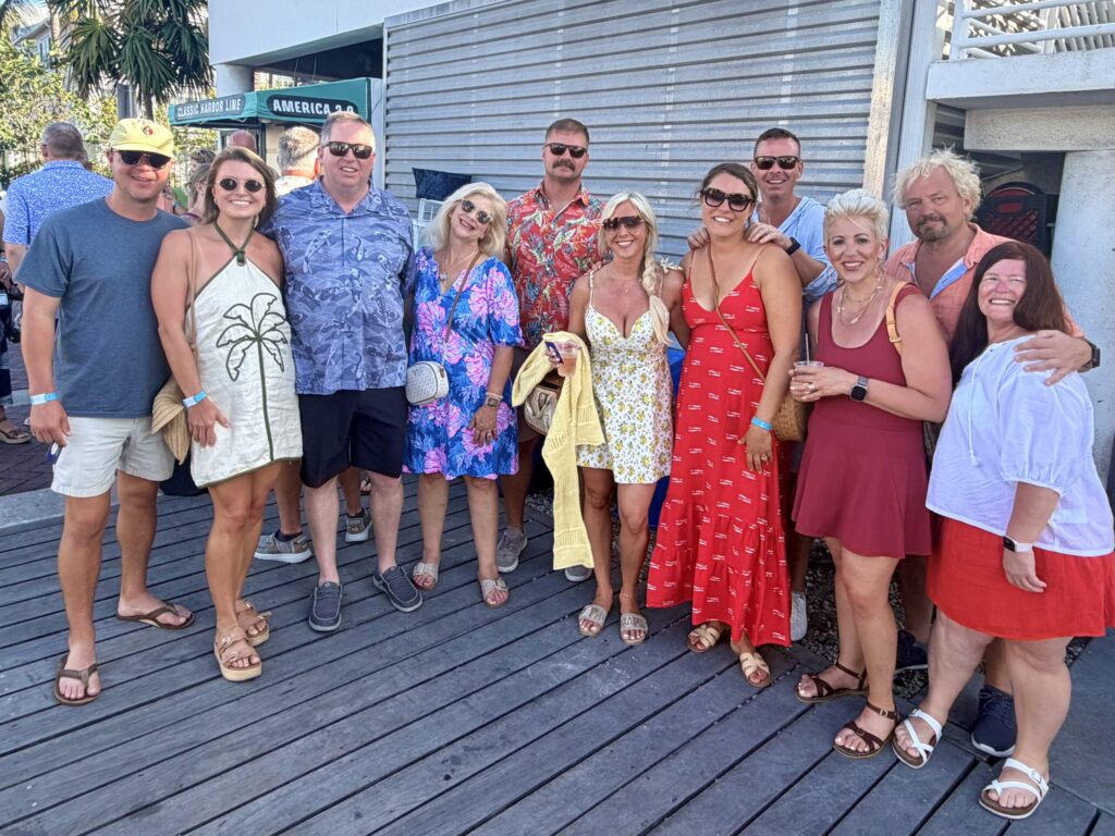 A group of men and women in vacation-wear cluster close on a wooden dock and smile.