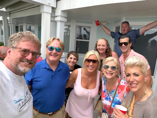 A group of men and women smile together on a covered patio, while another man raises his arms in celebration behind them.