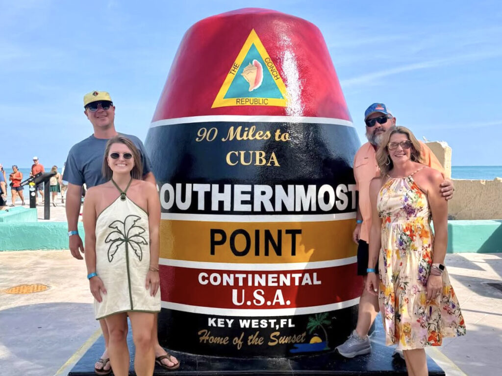 Two couples stand on either side of a giant painted buoy landmark that reads The Conch Republic / 90 miles to Cuba / Southernmost Point / Continental U.S.A. / Key West, Florida / Home of the Sunset.