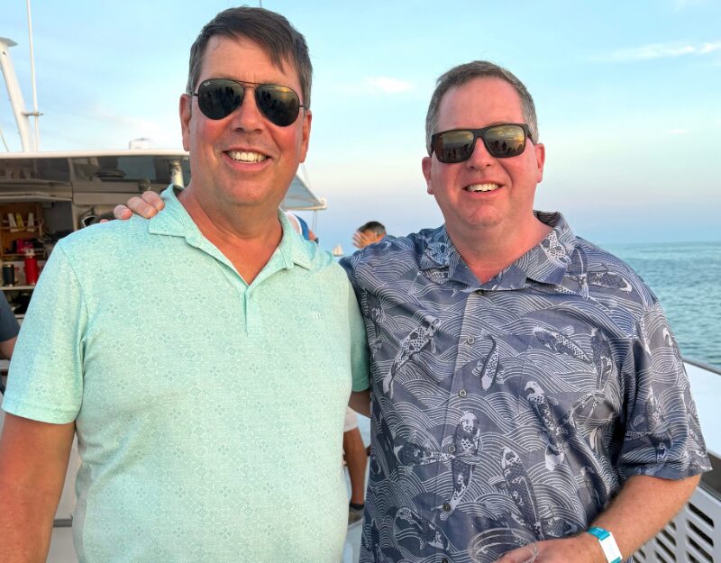 Two men wearing sunglasses and suntans smile with the ocean behind them.