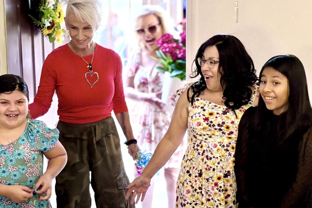 A blonde woman ushers a brunette woman and two young girls into a home through the front door, as the mother and daughters’ faces express shock and delight.