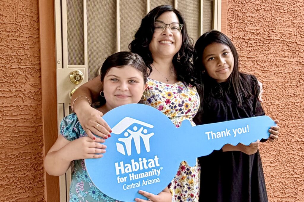 A woman wearing a floral dress with two young girls beside her, stands in front of a front door holding a blue sign shaped like a key that reads Habitat for Humanity Central Arizona | Thank You! 