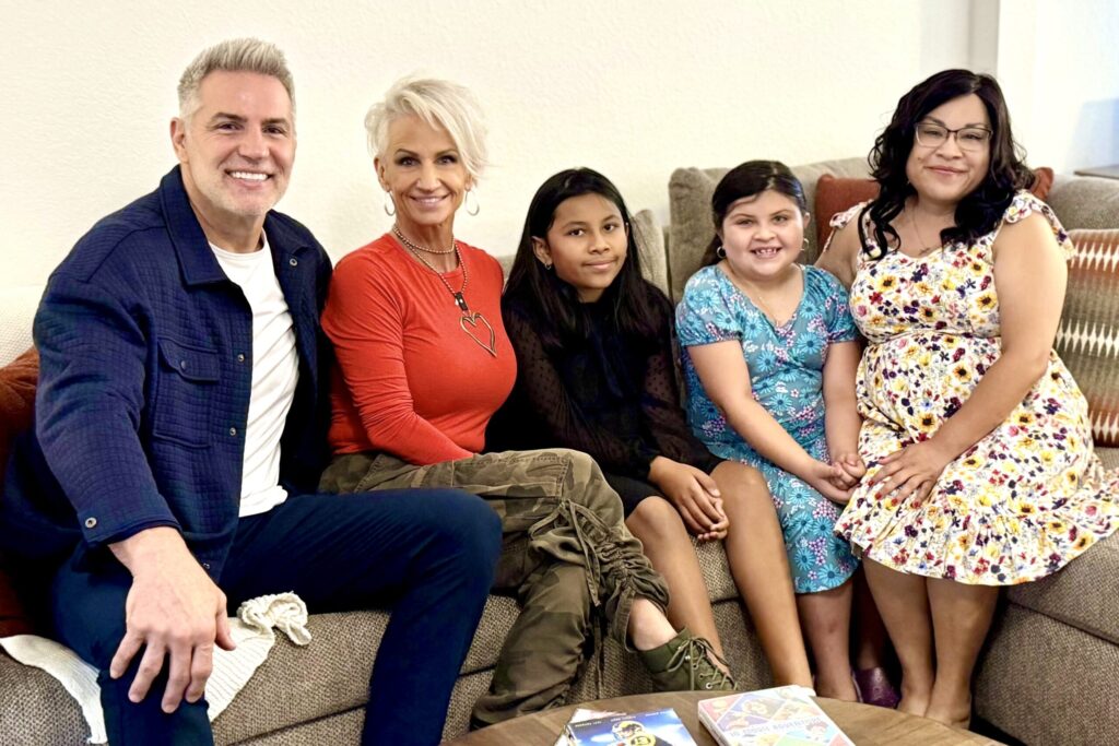 A bearded man and blonde woman sit on a sofa with a brunette woman and two young girls, all smiling.