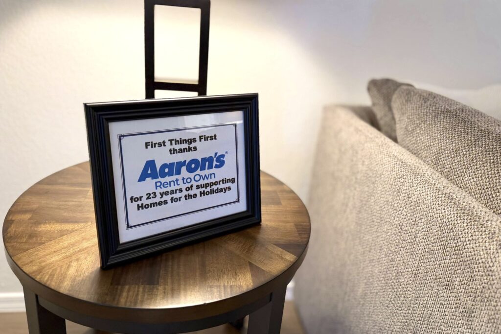 A blue and white framed sign reading “First Things First thanks Aaron’s Rent to Own for 23 years of supporting Homes for the Holidays” sits on a wooden end table beneath a lamp next to a sofa.