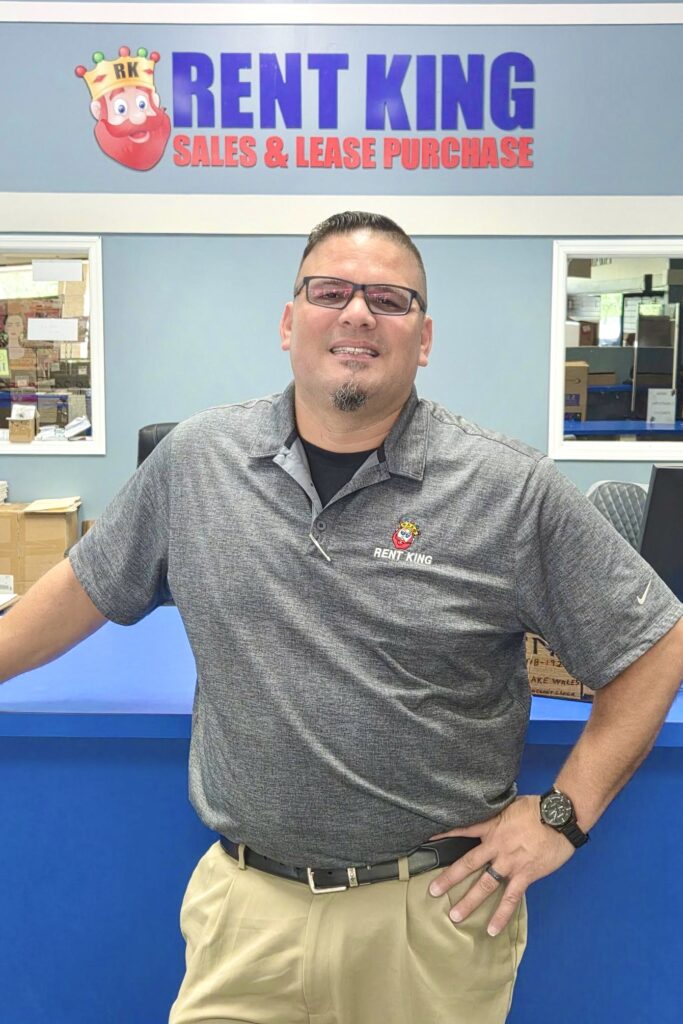 A man stands leaning against a counter and smiling, wearing spectacles, a Rent King sports shirt, and chinos. A sign behind him reads “Rent King | Sales & Lease Purchase.”