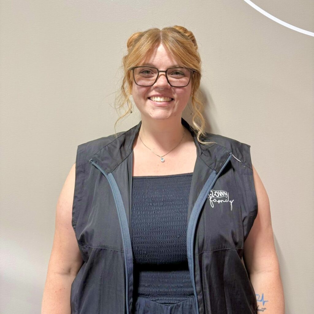 A young woman wearing spectacles, a navy blue dress, and a navy vest with the embroidered words “Rent-2-Own family” stands smiling.