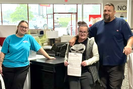 A young woman wearing a turquoise R2O tee stands with a man and woman holding up a rent-to-own contract in a showroom environment, all smiling.