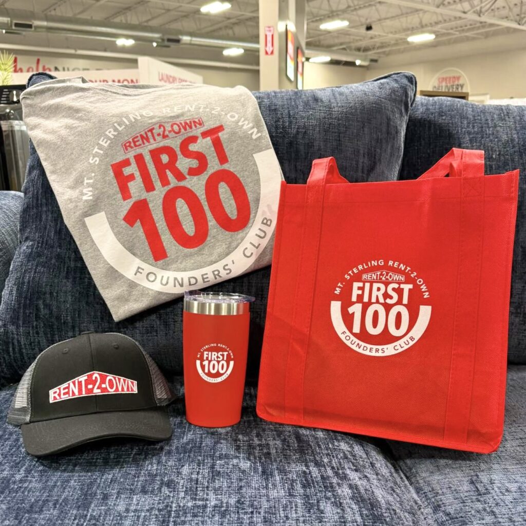 A grey tee, red tote and red travel cup – all branded with a circular logo reading “Mt. Sterling Rent-2-Own First 100 Founders’ Club” – and a black cap featuring the Rent-2-Own logo, are all displayed upon a dark blue sofa.