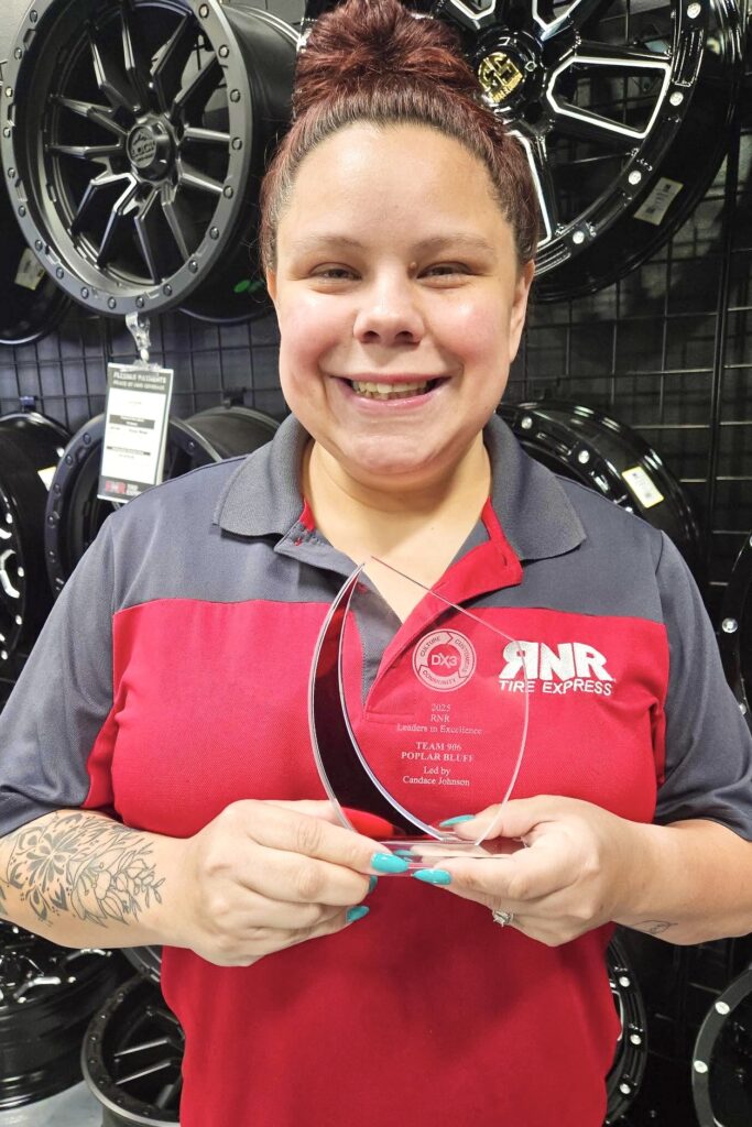 A young woman,
wearing a red and grey RNR Tire Express shirt, smiles
broadly as she holds up a crystal award, with a wall of tire rims hanging
behind her.