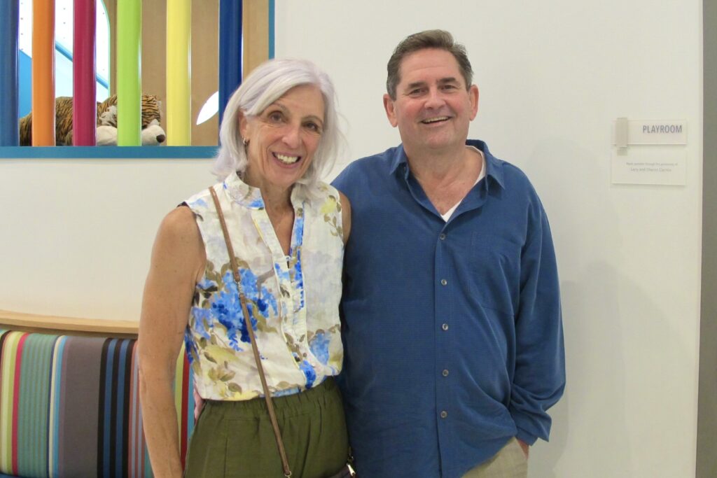 A woman with a floral blouse and a man with a blue shirt stand together smiling, with a wall plaque to their right reading “Playroom | Made possible through the generosity of Larry and Sharon Carrico.”