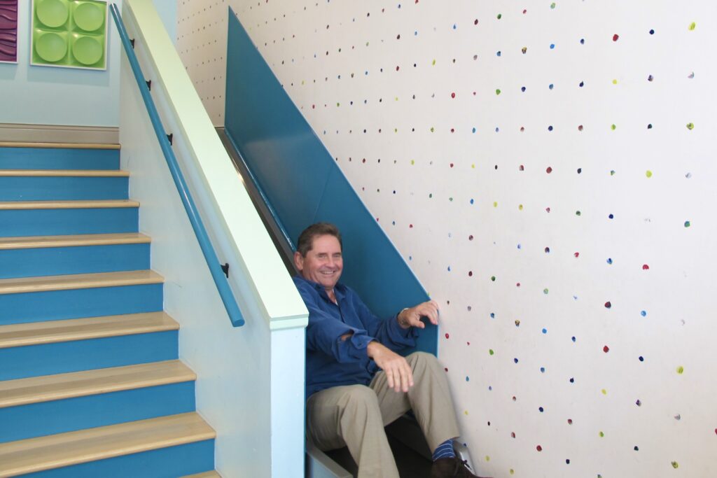 A middle-aged man wearing a blue shirt laughs as he’s awkwardly positioned at the bottom of a blue built-in slide.