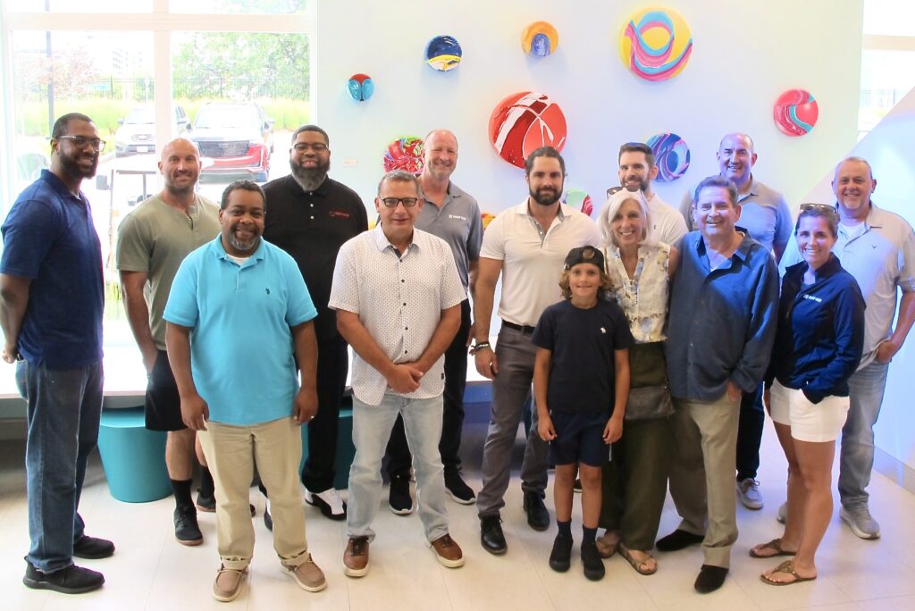 A small, diverse group of people stand smiling with bright windows and colorful artwork behind them.