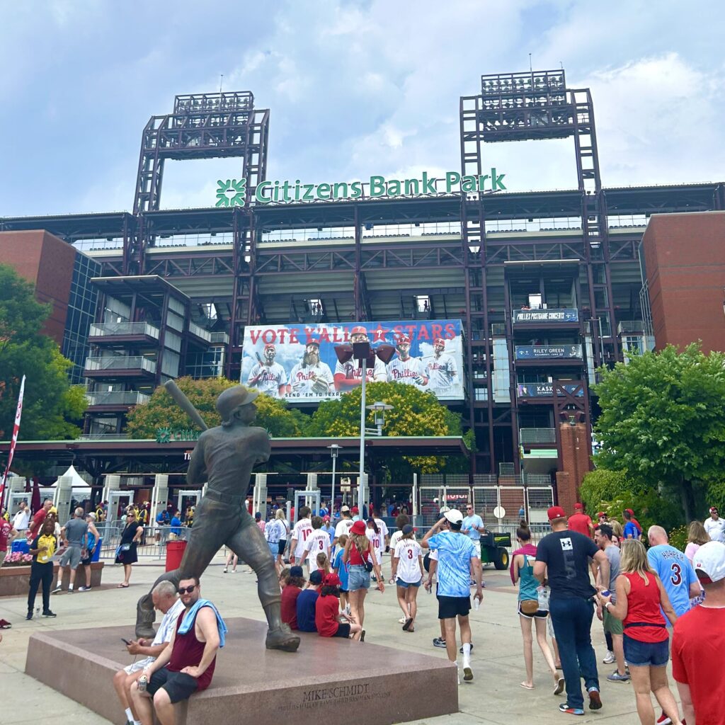 Dozens of people sit, stand, and walk around a two-story ballpark entrance topped with a Citizens Bank Park sign.