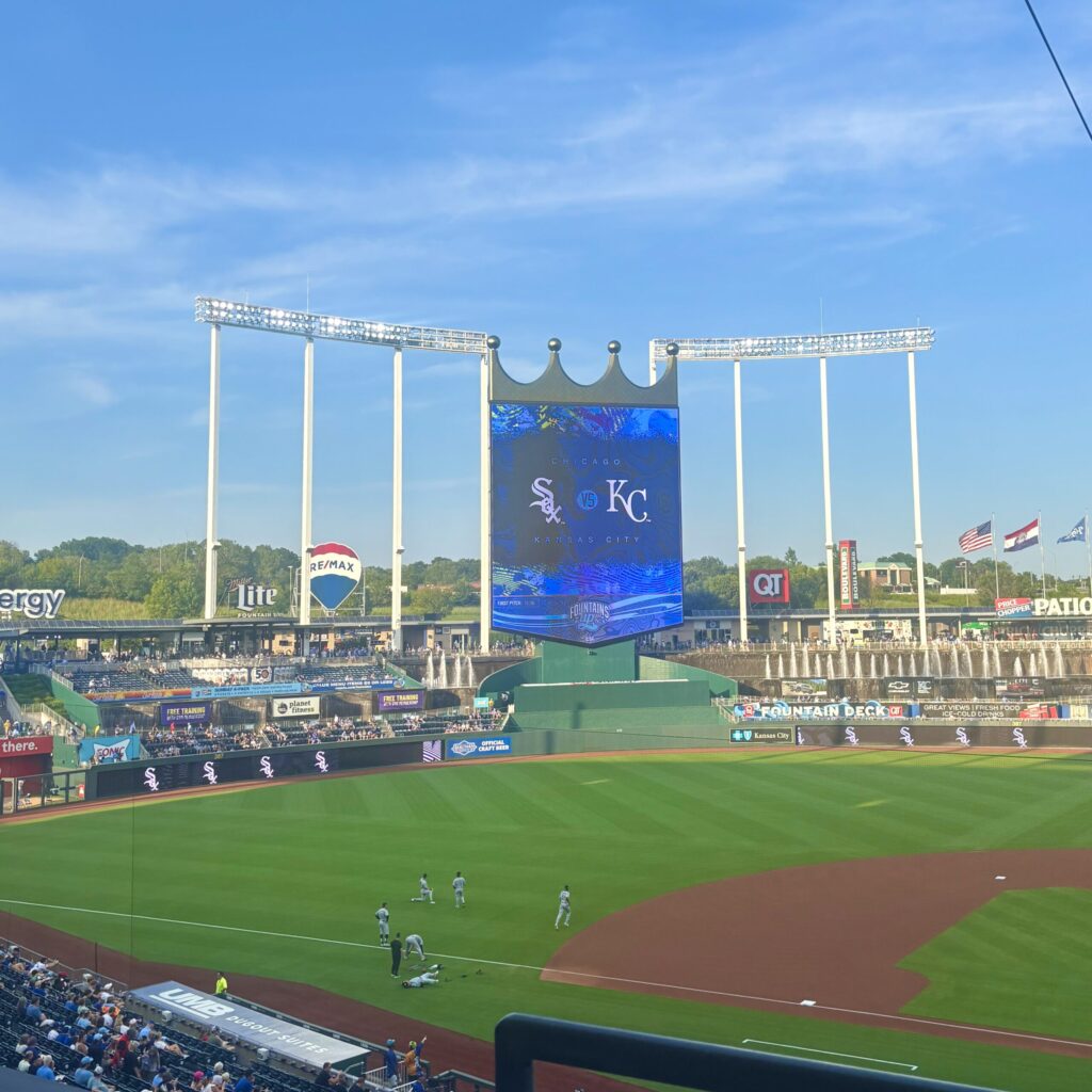 A jumbo screen topped with a gold crown shows a split screen of the Chicago White Sox logo and the Kansas City Royals logo showing over a bright green baseball field and a few players on a bright, clear day.