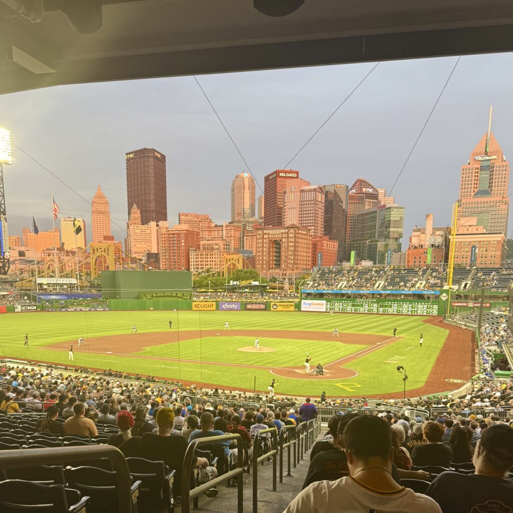 View from the crowd of a vibrant green baseball field with a full and dynamic city skyline behind it at dusk.