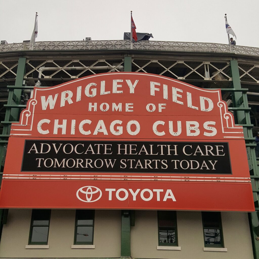 A large, red, vintage sign up on a big stone building reads Wrigley Field | Home of Chicago Cubs. Flags above the sign mark the top of the ballpark.