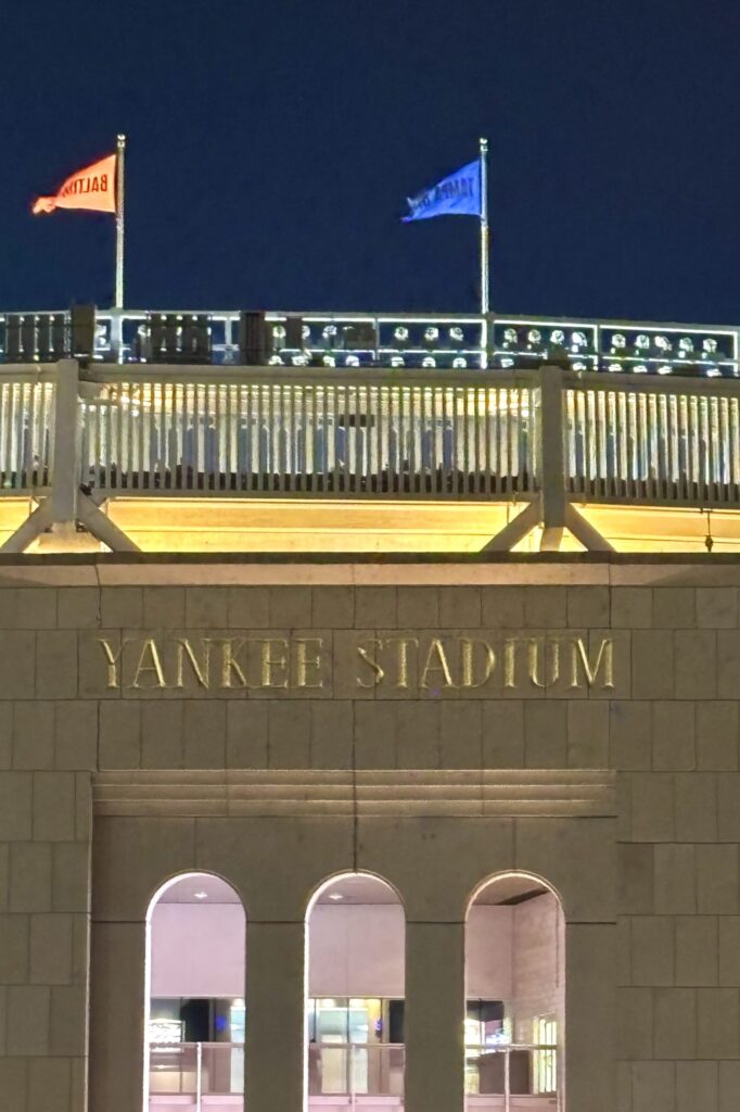 A stone building with three archways and a sign above them reading Yankee Stadium. Flags above the sign mark the top of the ballpark.
