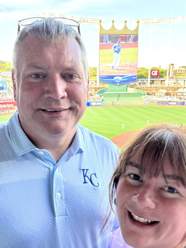 A couple smiles with a baseball park and jumbo screen behind them.