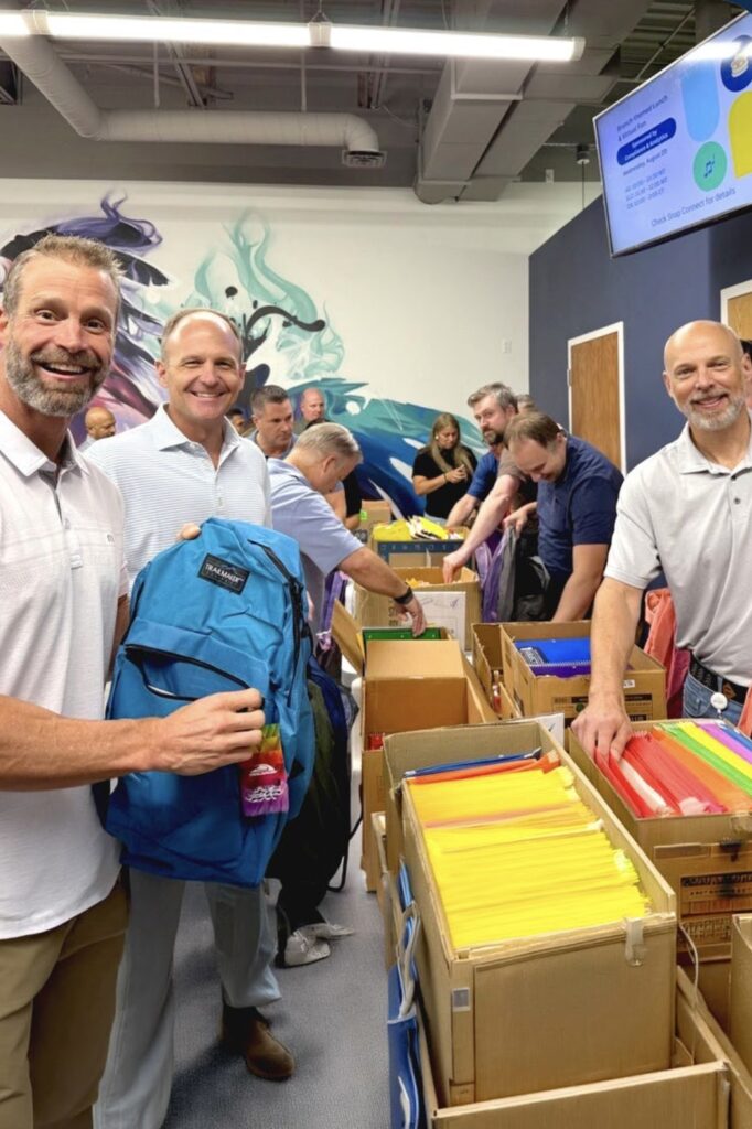 A group of adults stands around boxes full of colorful folders, working and smiling, with one man close to the camera holds up a bright blue backpack.