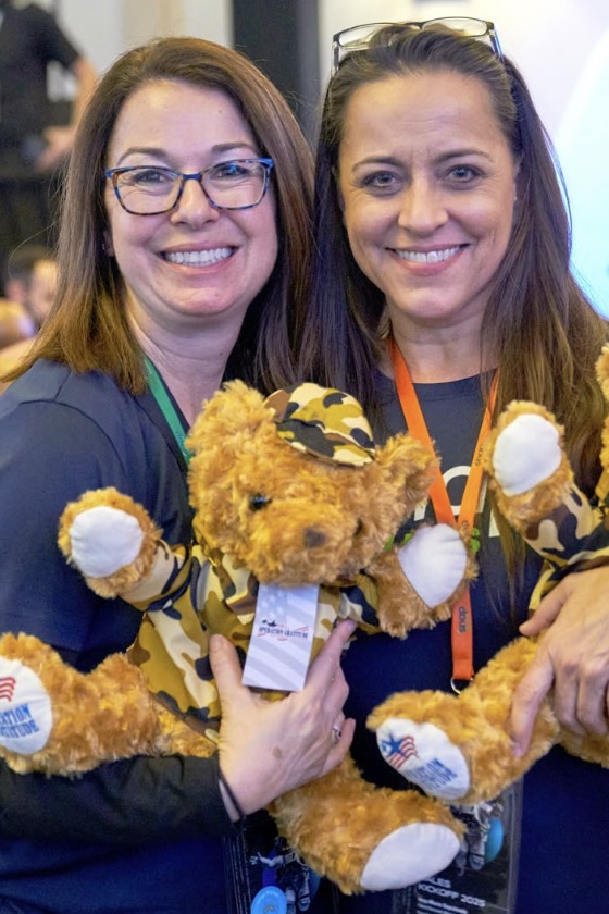 Two women stand together smiling, each holding a brown, fuzzy teddy bear wearing a camouflage hat and jacket, with “Operation Gratitude” embroidered onto one foot.