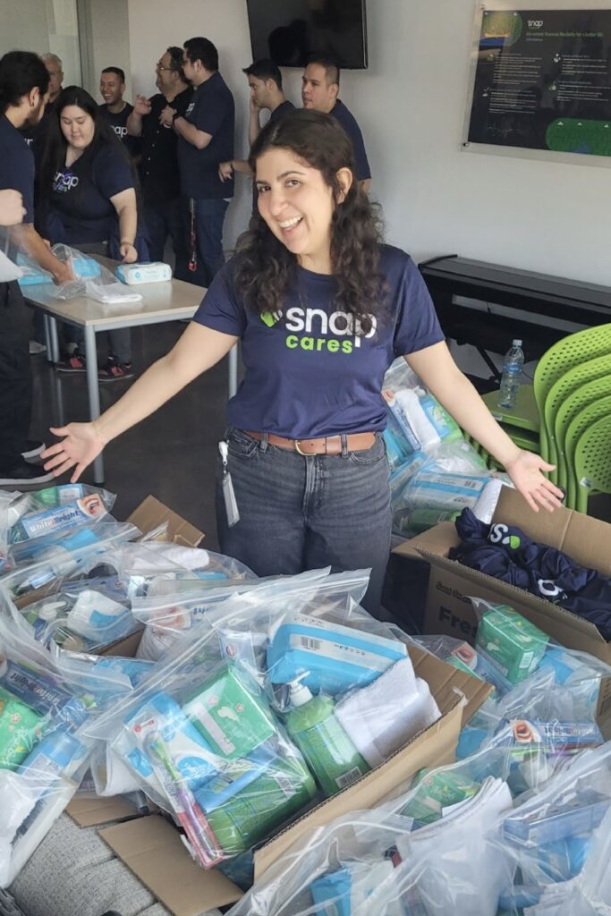 A brunette woman wearing a navy-blue tee with the “Snap Cares” logo smiles as she shows off dozens of storage bags filled with personal hygiene items; other volunteers continue working behind her.