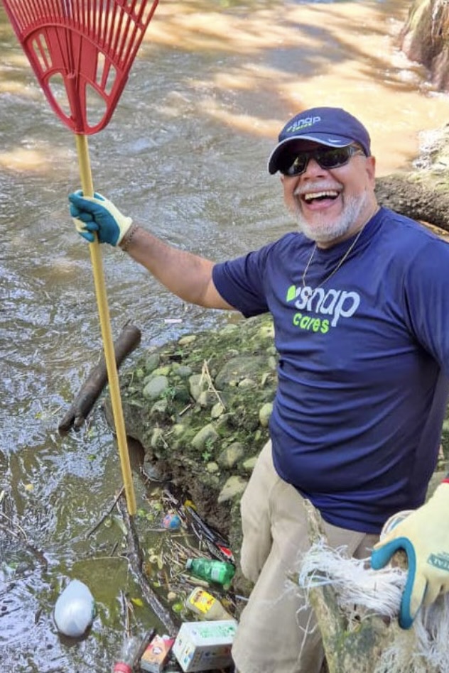 A smiling bearded man wears a navy-blue tee with the “Snap Cares” logo and holds a bright red rake as he helps clean up garbage from a waterway.