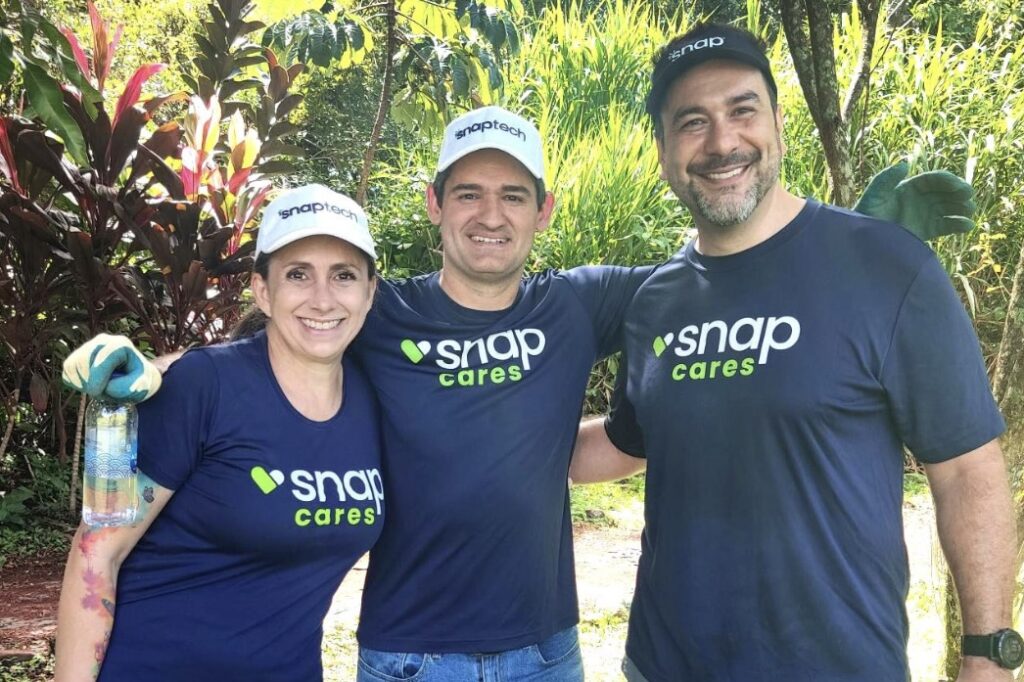Two men and a woman wearing navy-blue tees with “Snap Cares” logos, Snap baseball caps, and gardening gloves stand together smiling, with lush greenery behind them.