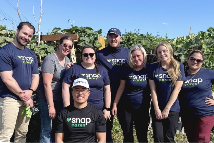 A group of men and women wearing navy-blue tees with “Snap Cares” logos standing together smiling, with tall green plants behind them.