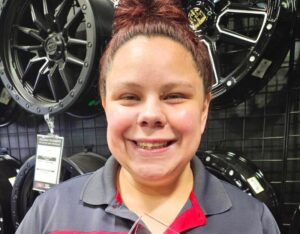 A young woman, wearing a red and grey RNR Tire Express shirt, smiles broadly as she holds up a crystal award, with a wall of tire rims hanging behind her.