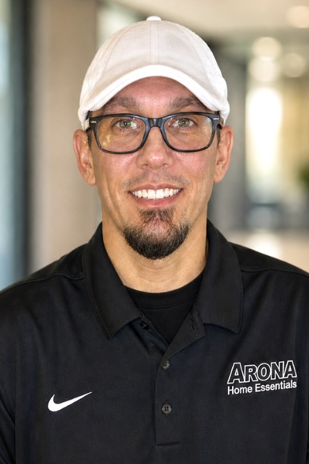 A man with black eyeglasses and a goatee smiles while wearing a white cap and black polo shirt with a white Arona Home Essentials logo on it.