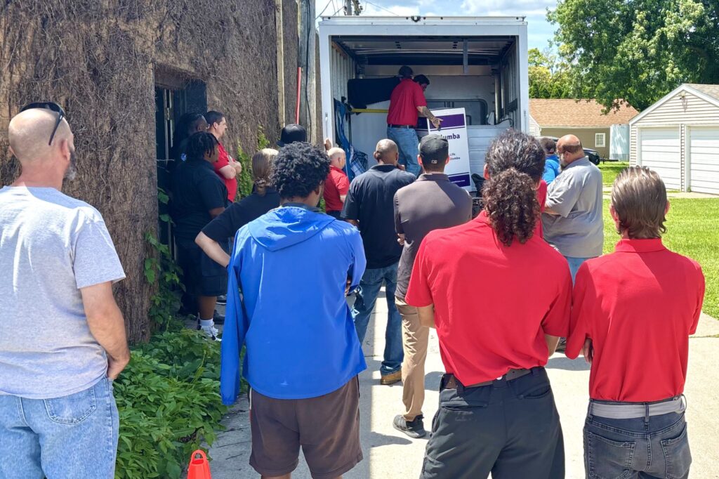 A crowd faces the open back of a delivery truck, watching two men working inside of it.