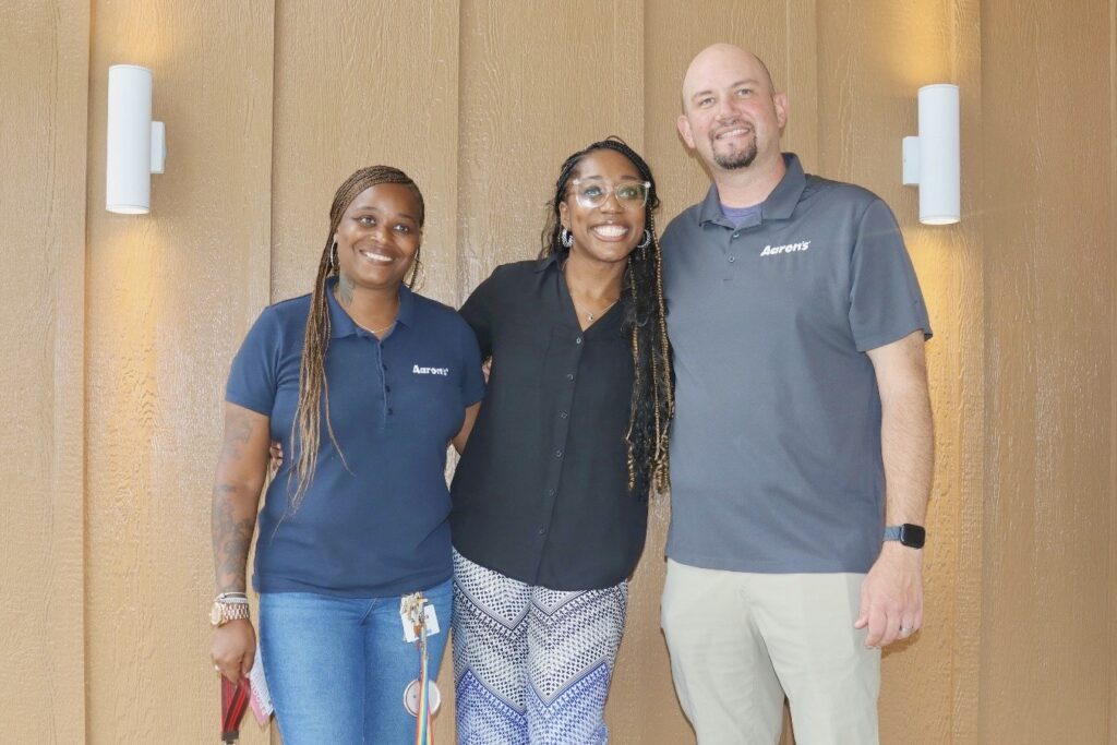 A man and woman wearing navy blue polo shirts with Aaron’s logos stand on either side of a third woman, all smiling.