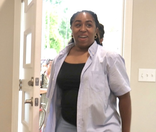 A casually dressed woman smiles broadly as she walks through an open door, holding a gift bag that reads ‘Best Day Ever.’
